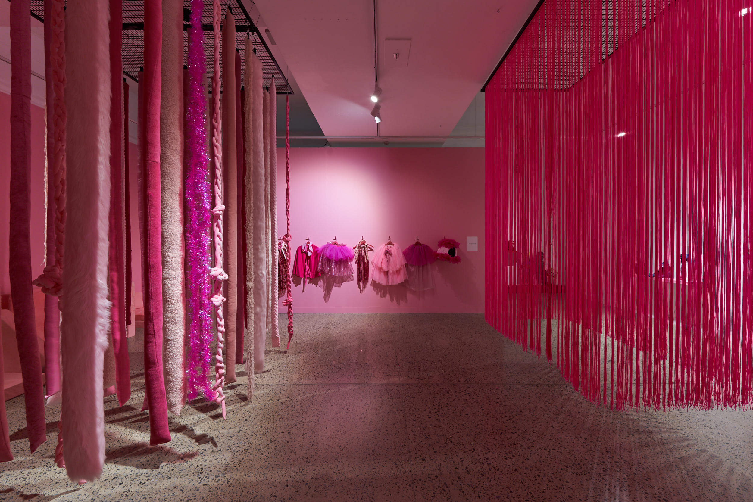 Installation view of a contemporary art exhibition dominated by shades of pink. Long, textured fabric and yarn pieces hang from the ceiling on the left, while a dense curtain of pink cords creates a partition on the right. Pink garments and sculptural textile works are displayed on the back wall in a softly lit gallery with a terrazzo floor.